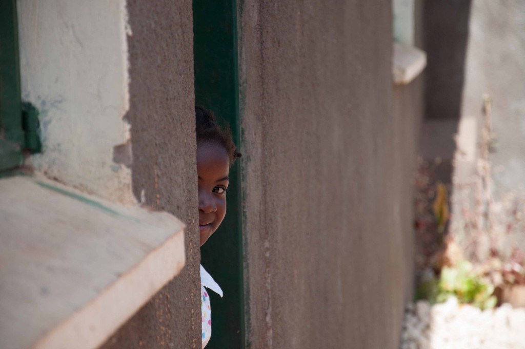 A young girl peeks around the door of a house on the school property.